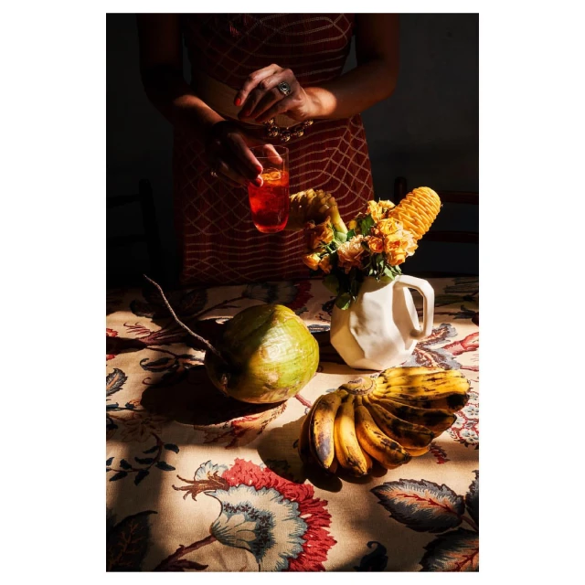 Still life photograph featuring a woman holding a cocktail alongside fruit, flowers, and a patterned tablecloth, capturing a warm, inviting atmosphere.