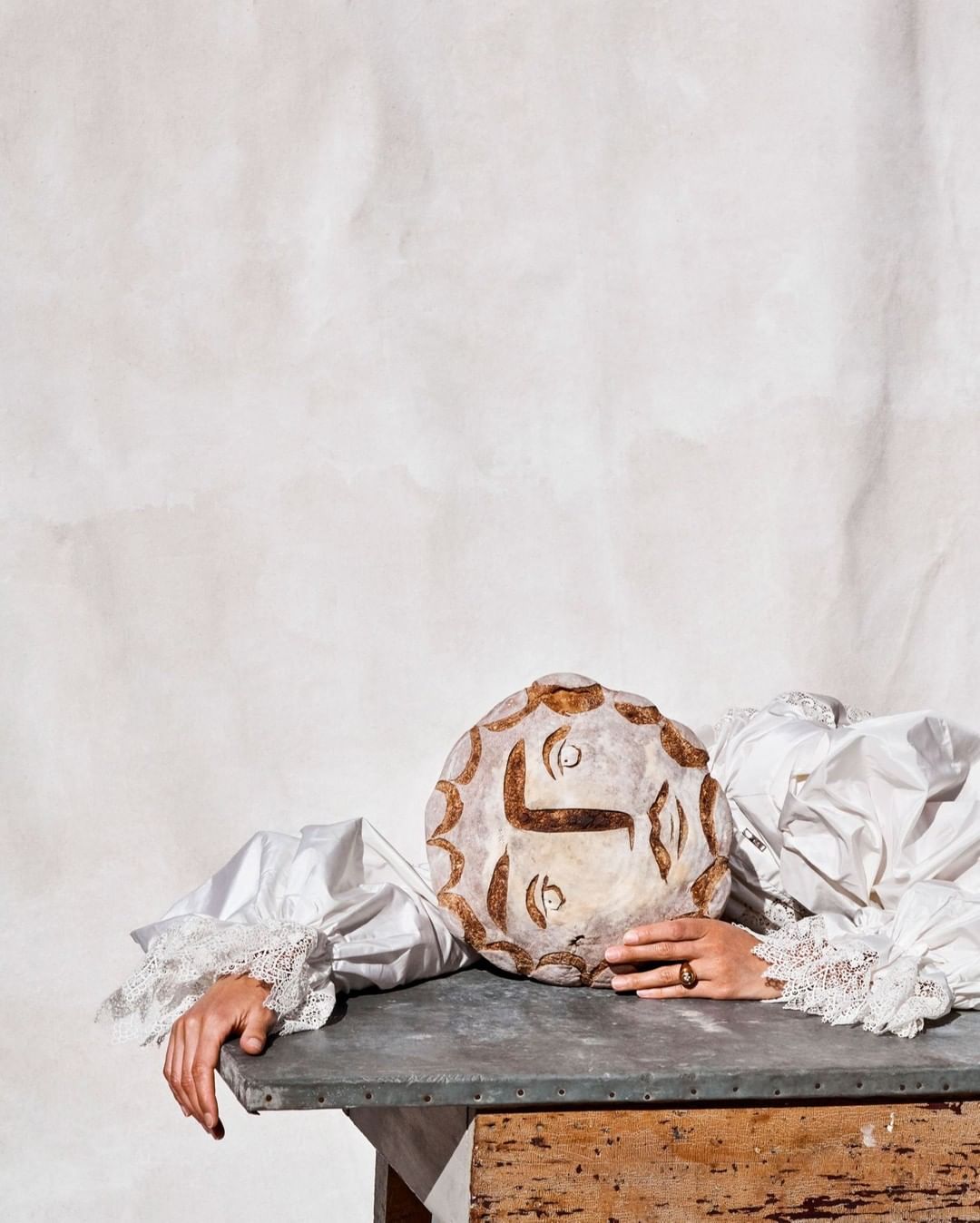Still life of an artistically scored loaf of bread on a gray surface with a person's hands and lace-cuffed sleeves visible.