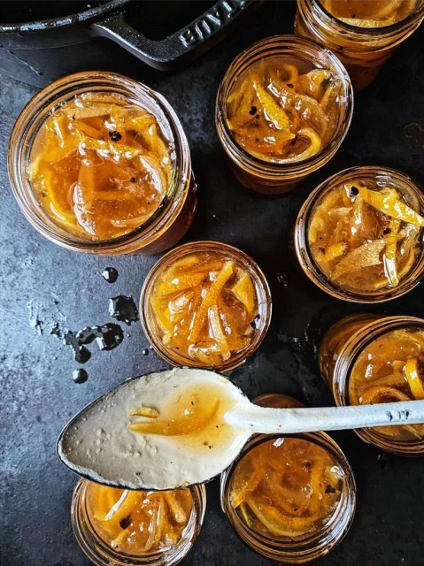 Overhead shot of mason jars filled with orange marmalade, sitting on a dark surface, with a spoonful of marmalade in the foreground.