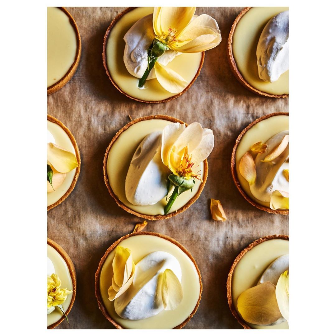 Overhead shot of nine tartlets arranged on parchment paper, topped with light yellow custard, white cream, and yellow flower petals.