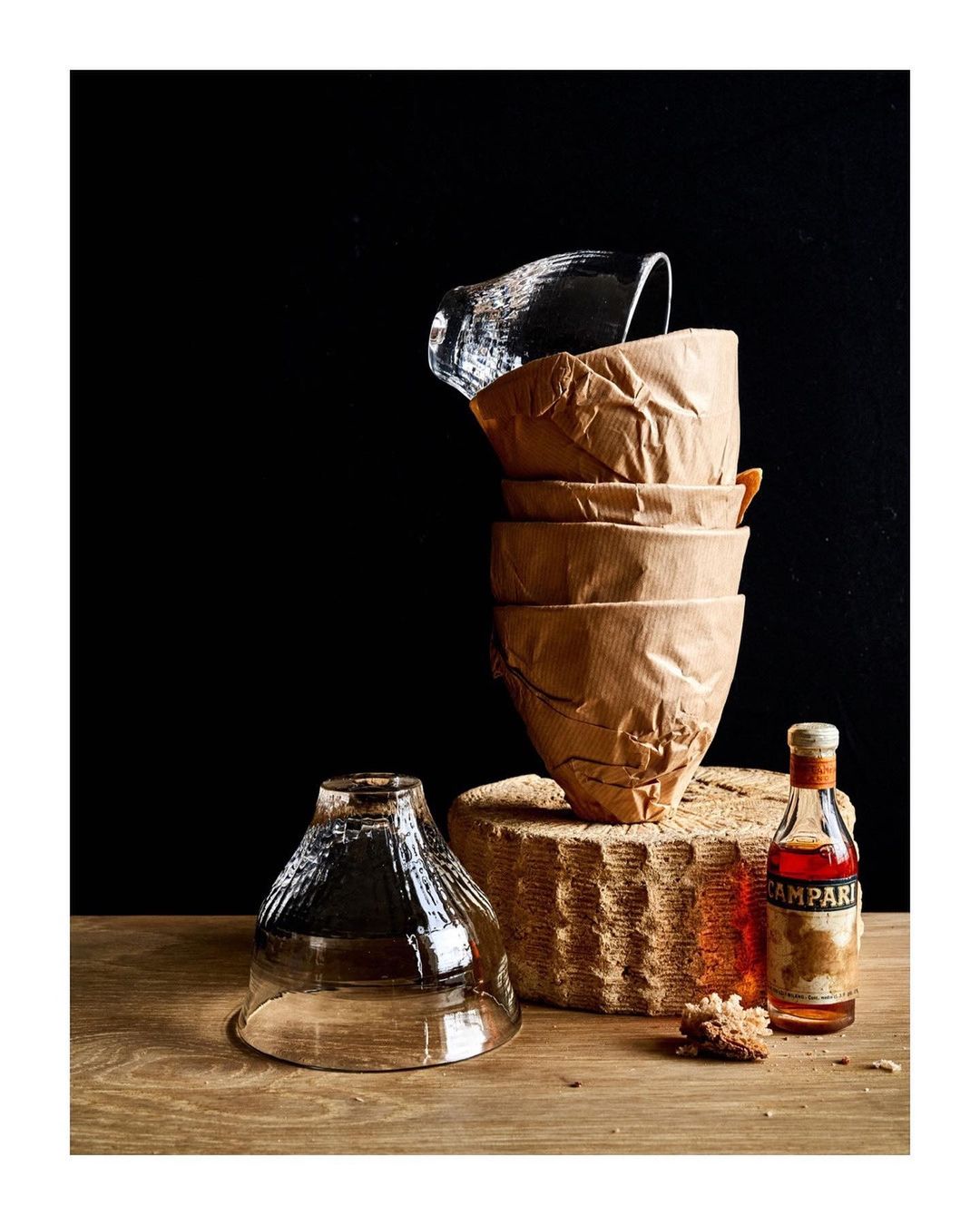 Still life shot with stacked glassware, paper cups, and a Campari bottle on a wooden surface against a dark background.