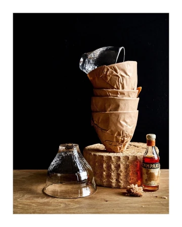 Still life shot with stacked glassware, paper cups, and a Campari bottle on a wooden surface against a dark background.
