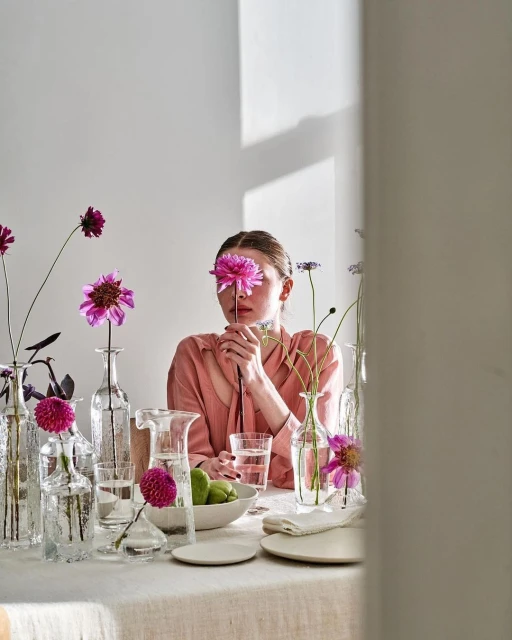 Woman with a pink flower over her eye sitting at a table with flower-filled vases, plates, a bowl of fruit, and a glass of water.
