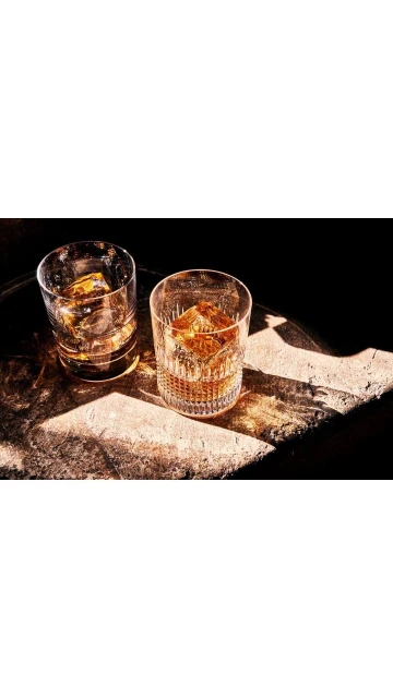 Two glasses of whiskey with ice sit on a stone surface with dark background, illuminated by a warm light source, studio shot.