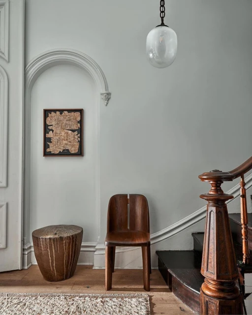Interior design with wooden chair, rustic side table, artwork, and staircase against a light gray wall with globe pendant light.