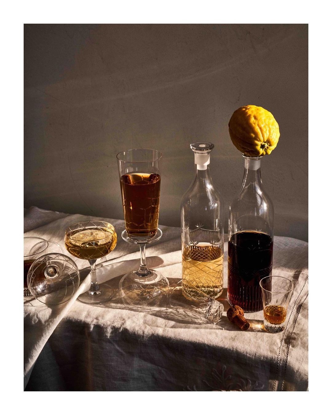 Still life of glass bottles and stemmed glasses filled with drinks, alongside a lemon on a linen-covered table in warm light.