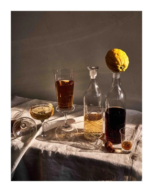 Still life of glass bottles and stemmed glasses filled with drinks, alongside a lemon on a linen-covered table in warm light.