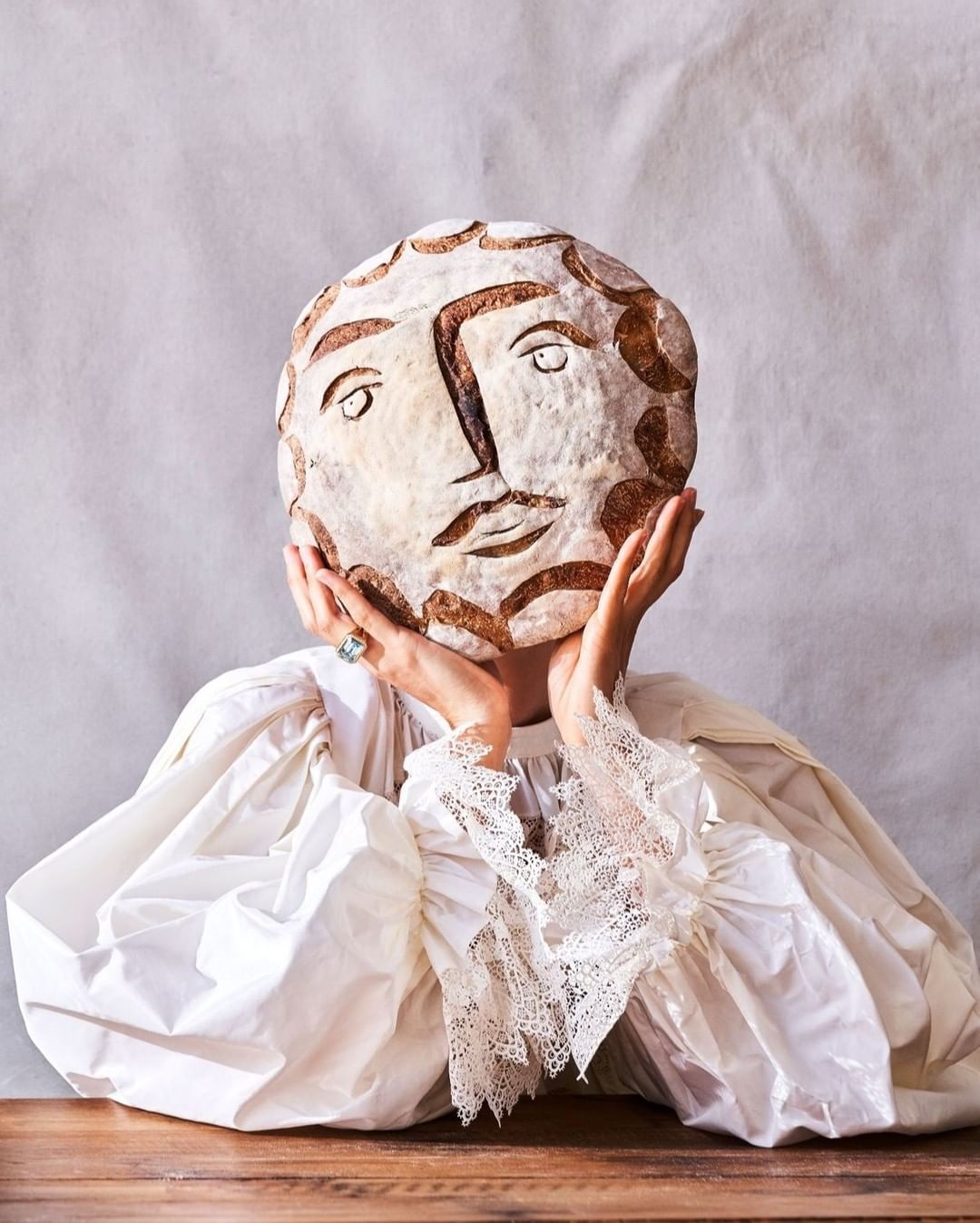 Woman in ruffled sleeves holds a bread loaf with a face carved into it, obscuring her face against a white backdrop on a wood table.