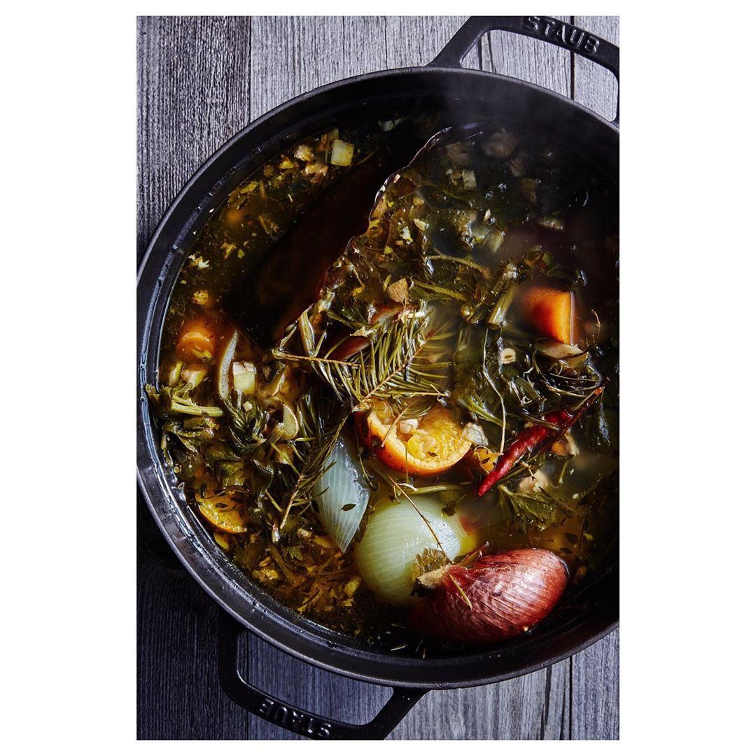 Overhead shot of vegetable stock cooking in black Staub pot on gray wood background; including onions, carrots, herbs, and spices.