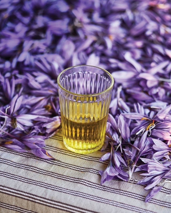 A glass of clear liquid sits surrounded by scattered purple flower petals on a striped tablecloth, capturing a natural, serene scene.