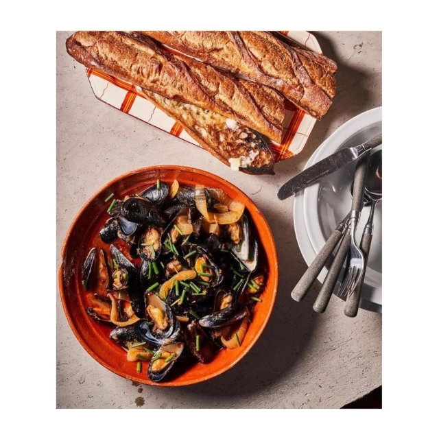 Overhead shot of a plate of steamed mussels with herbs, alongside a sliced baguette and silverware on a neutral-colored surface.