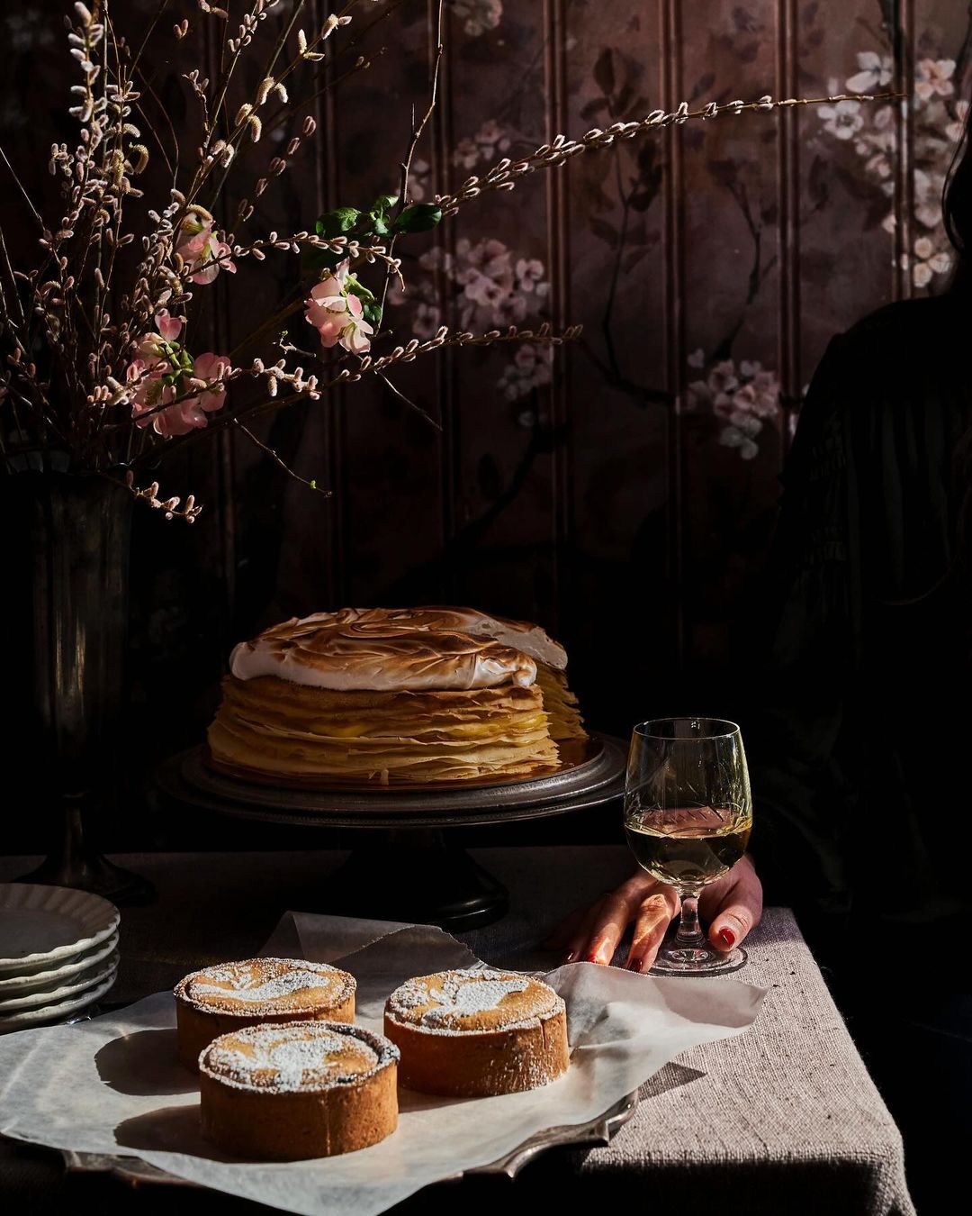 Overhead shot of dessert table with crepe cake, tartlets, wine, and flower arrangement against a dark floral wallpaper backdrop, indoors.