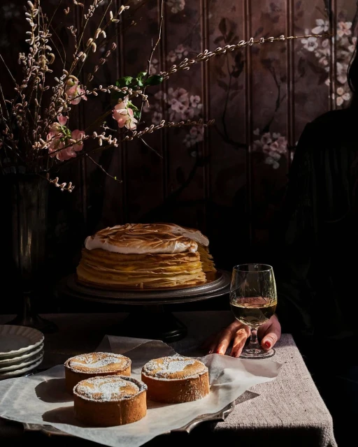 Overhead shot of dessert table with crepe cake, tartlets, wine, and flower arrangement against a dark floral wallpaper backdrop, indoors.