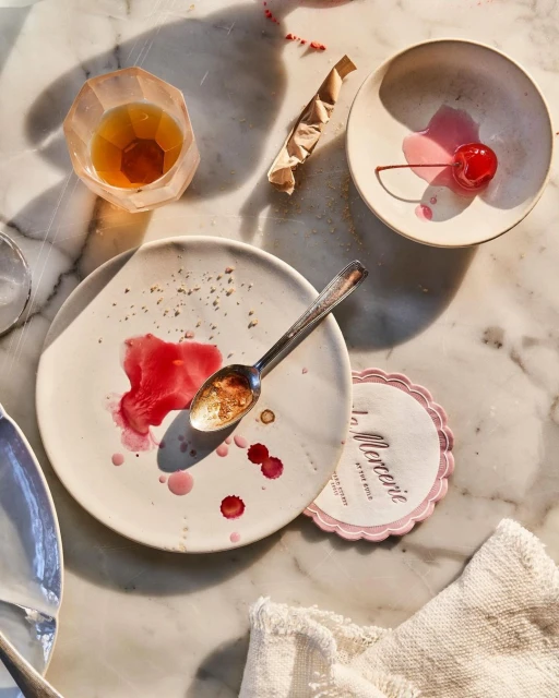 Overhead shot of a messy table with dirty plates, silverware, a half-eaten cherry, a cocktail, and a coaster with La Mercerie text.