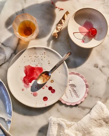 Overhead shot of a messy table with dirty plates, silverware, a half-eaten cherry, a cocktail, and a coaster with La Mercerie text.