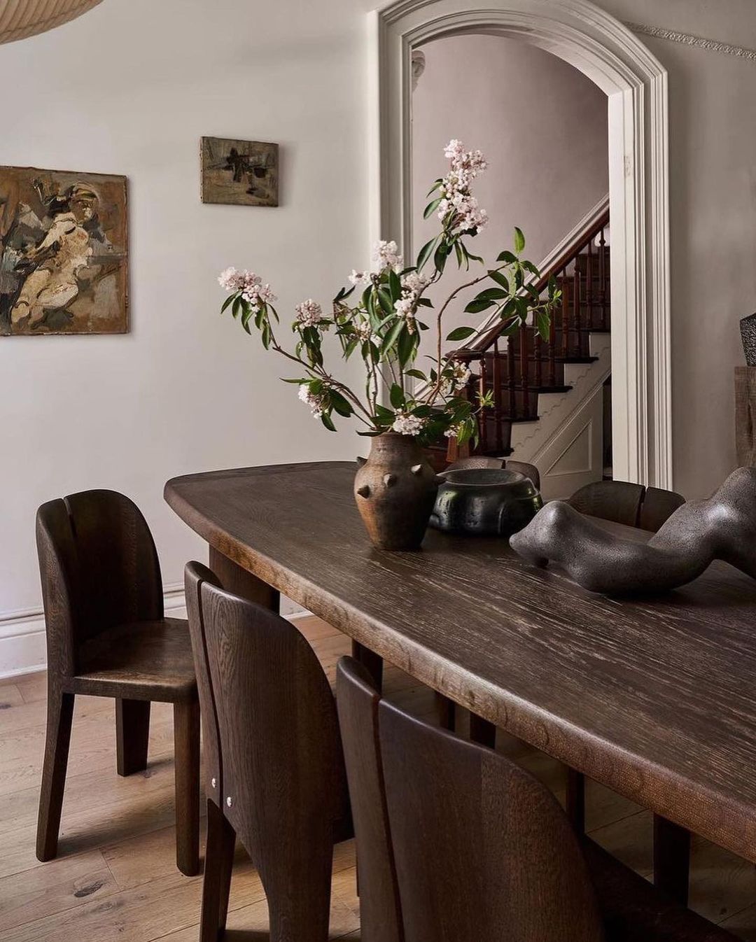 Dining room with dark wood table, chairs, and brown vase with flowers, accented by paintings on the wall and stairs in the background.