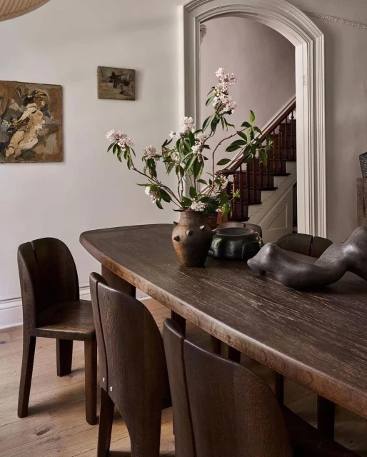 Dining room with dark wood table, chairs, and brown vase with flowers, accented by paintings on the wall and stairs in the background.