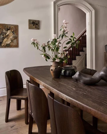 Dining room with dark wood table, chairs, and brown vase with flowers, accented by paintings on the wall and stairs in the background.