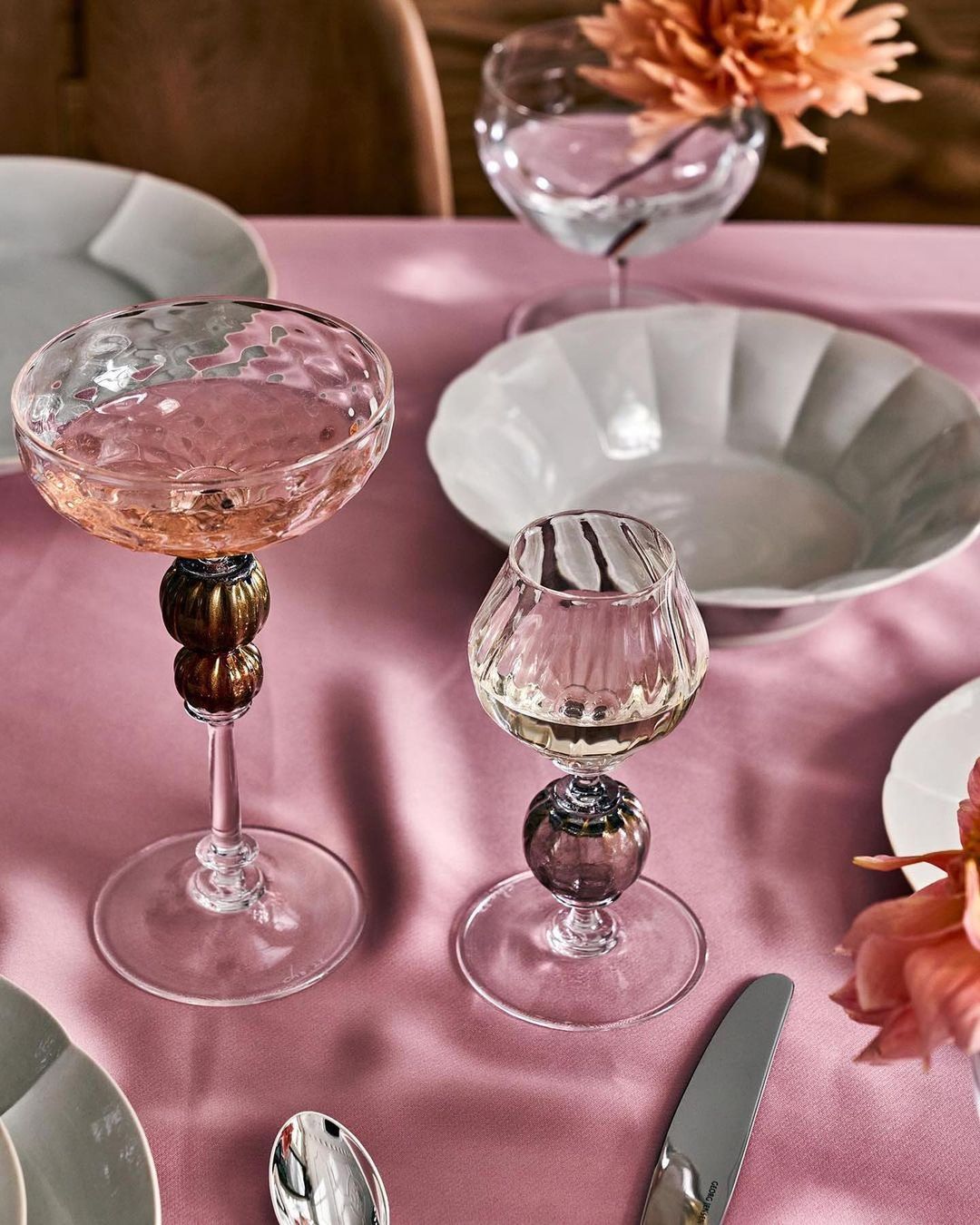 Elegant table setting with two decorative stemmed glasses, white plates, silverware, and a pink tablecloth create a refined atmosphere.