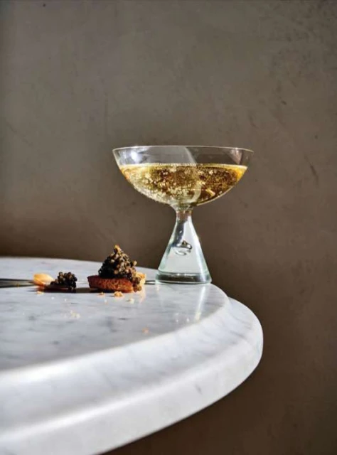 A glass of sparkling wine next to a canapé topped with caviar sits on a white marble table against a neutral backdrop.