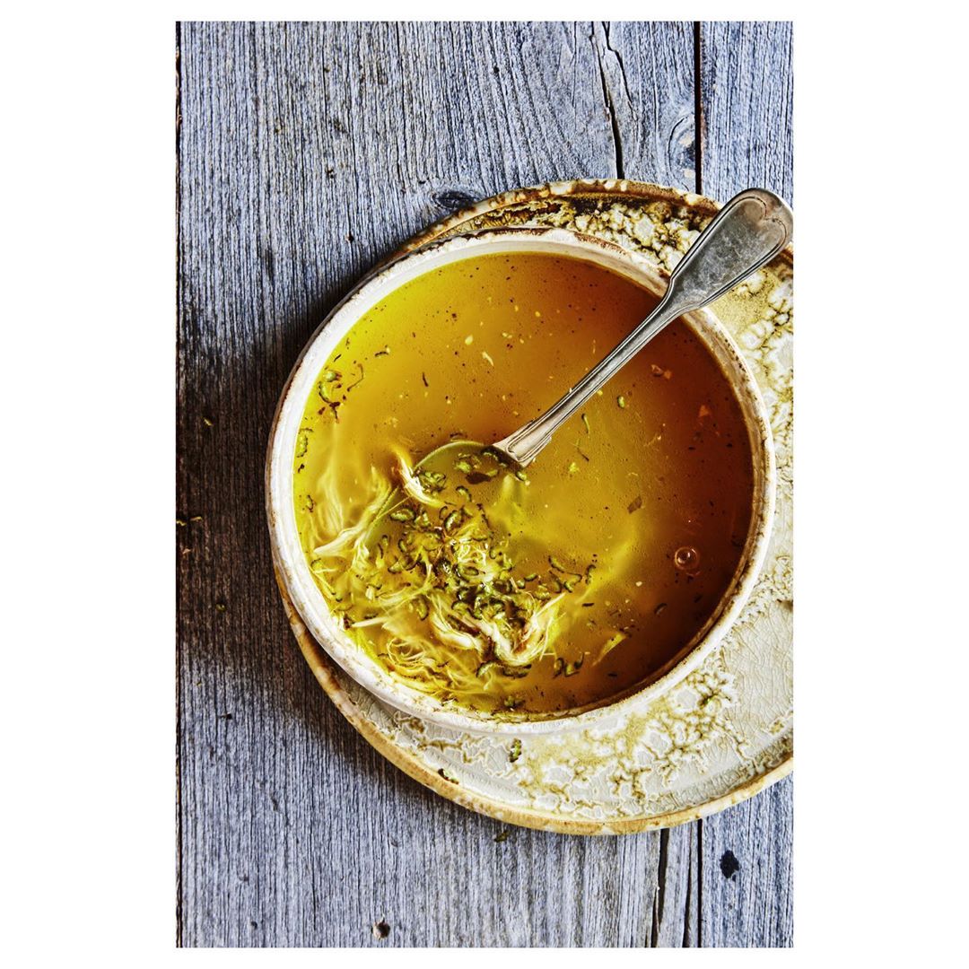 Overhead view of a bowl of chicken noodle soup with a spoon, placed on a rustic wooden surface with a speckled plate.