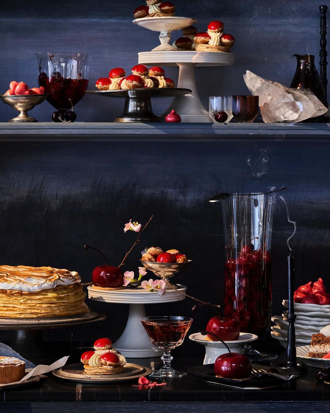 Still life of cherry-topped pastries, cakes, and beverages arranged on tiered stands against a dark background, evoking a moody atmosphere.
