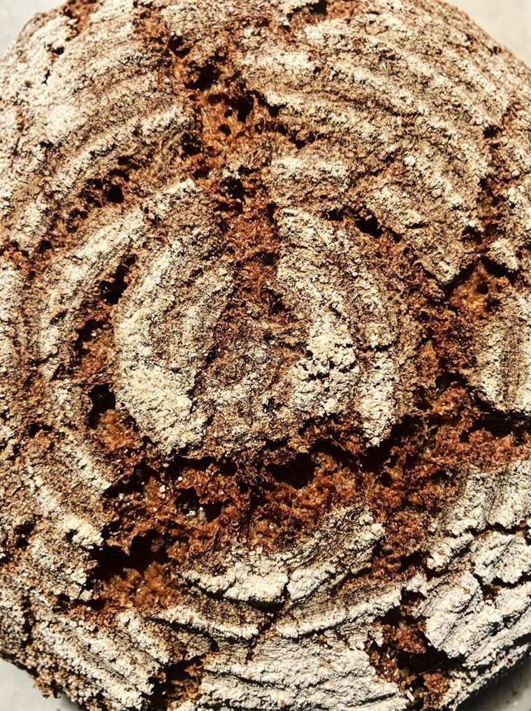 Close-up of freshly baked round bread loaf, showing its textured crust with concentric rings of flour against a dark brown background.