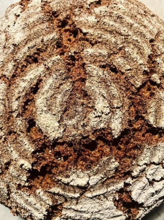 Close-up of freshly baked round bread loaf, showing its textured crust with concentric rings of flour against a dark brown background.