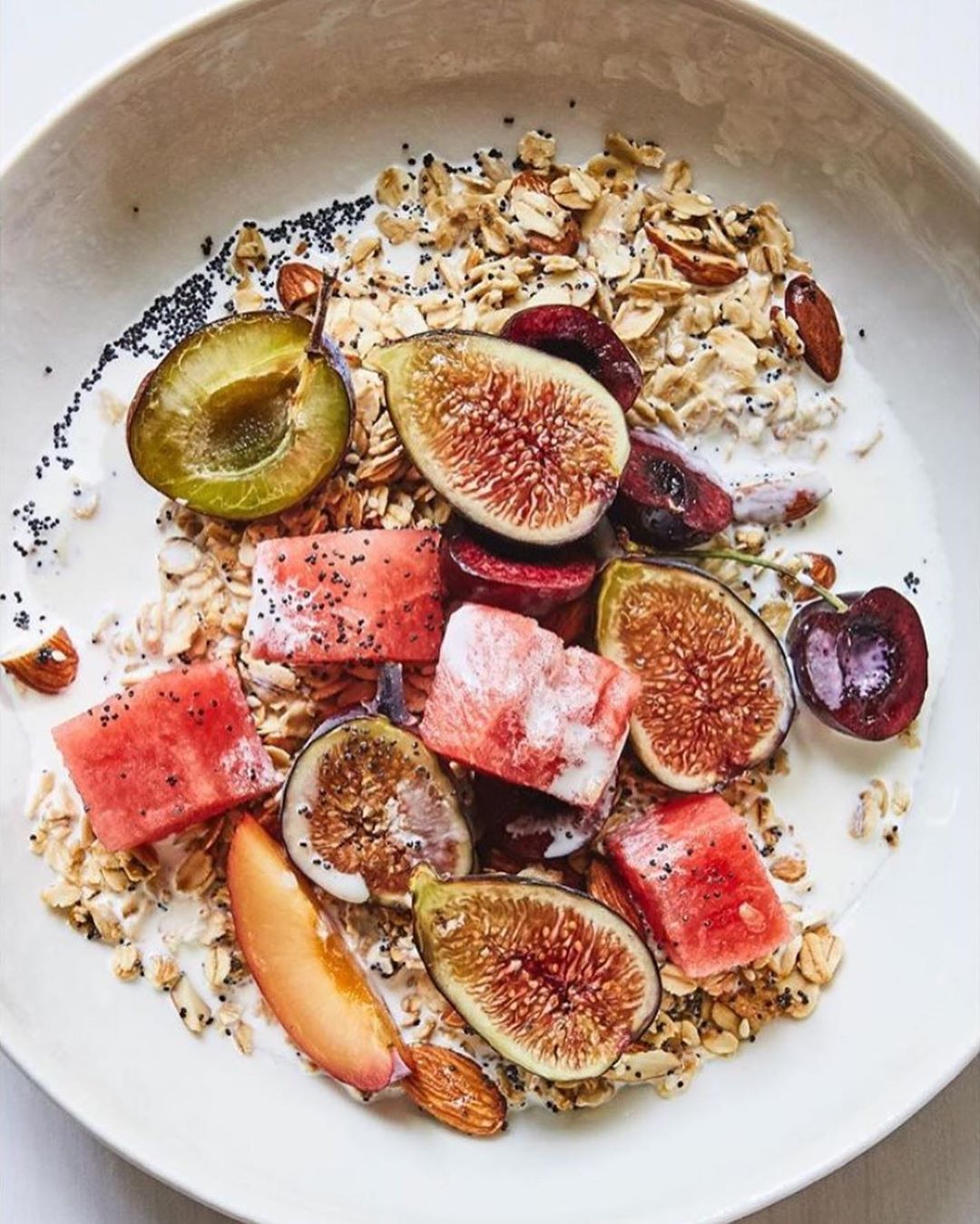 Overhead shot of a white bowl filled with yogurt, granola, figs, watermelon, cherries, and plums arranged on a white surface.