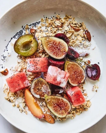 Overhead shot of a white bowl filled with yogurt, granola, figs, watermelon, cherries, and plums arranged on a white surface.