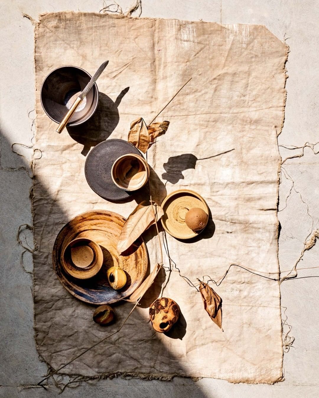 Overhead shot of bowls, plates, flatware and dried botanicals arranged on a tan textile with shadows and natural light creating a calming scene.