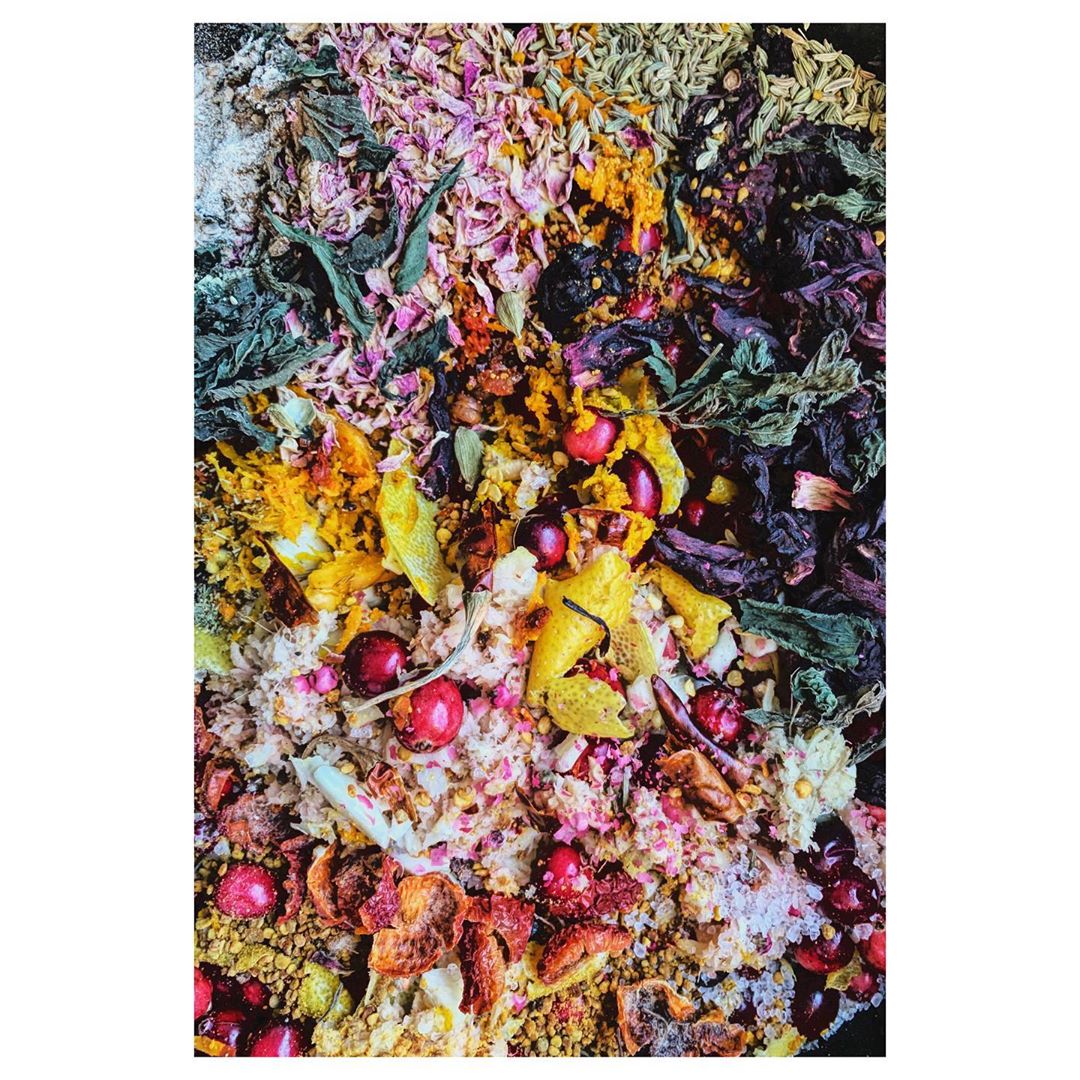 Overhead close-up shows various dried herbs, spices, and flower petals in vibrant colors: rose, yellow, green, and red, on a dark surface.