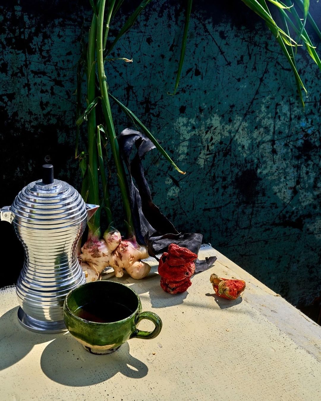 Still life of a silver moka pot and green cup with ginger root and sprouts, set against a textured teal and black background.