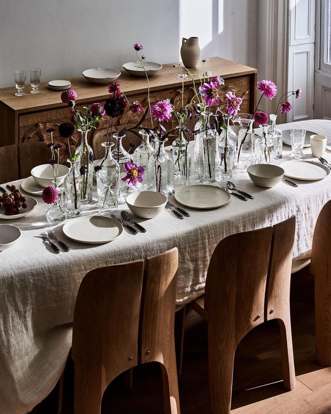 Formal dining table set with white linen, plates, bowls, silverware, clear vases with purple flowers, wood sideboard, and wood chairs.