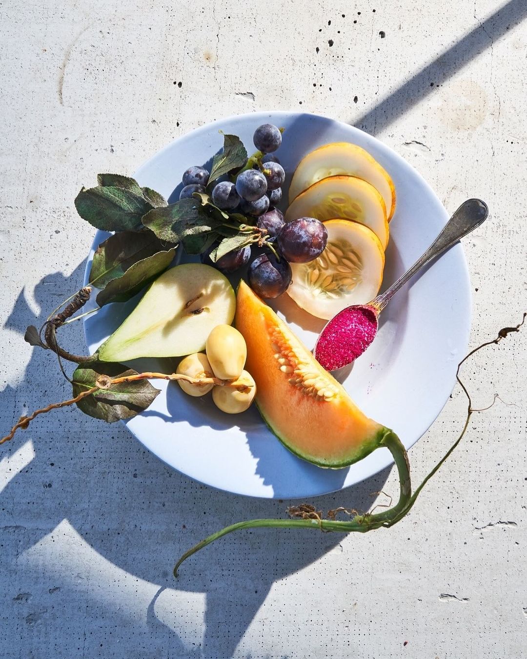 Overhead shot of a plate of food with a pear half, grapes, cantaloupe, honeydew, and a spoon with red jam, sitting on a concrete surface.