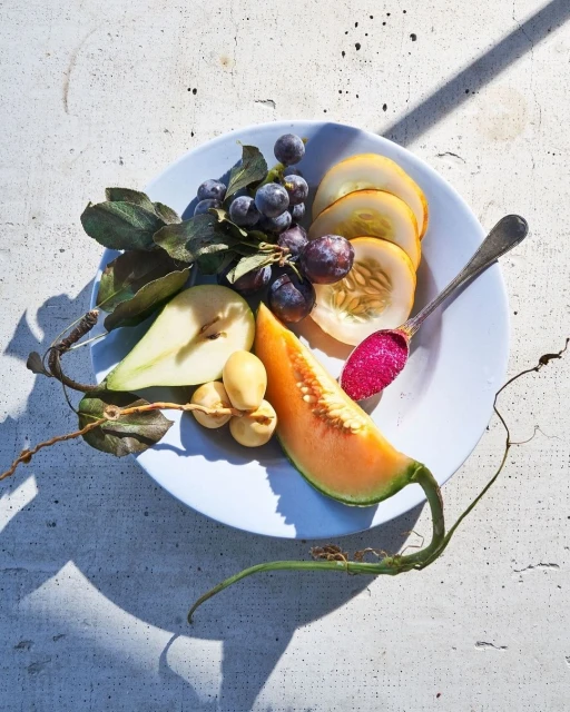 Overhead shot of a plate of food with a pear half, grapes, cantaloupe, honeydew, and a spoon with red jam, sitting on a concrete surface.