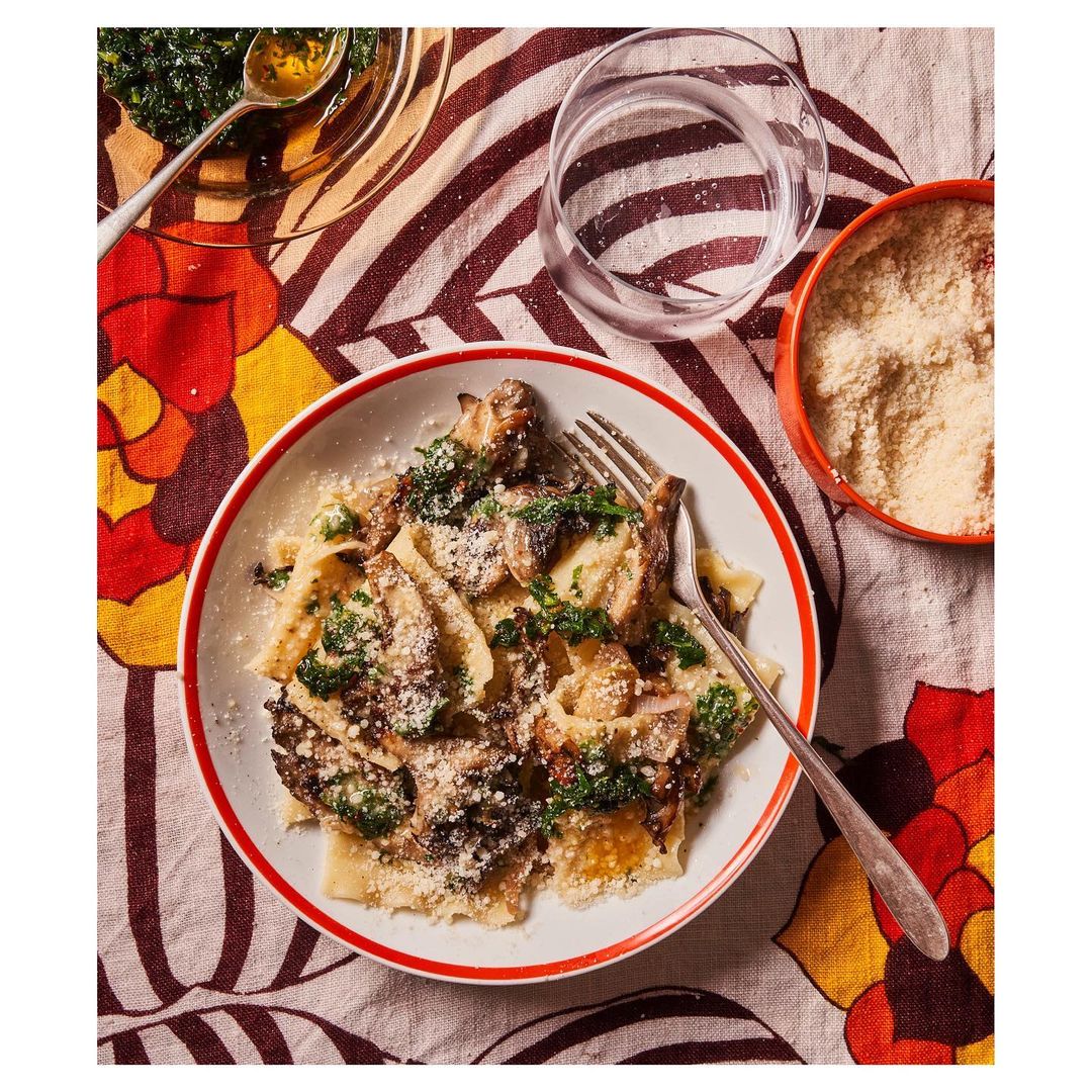 Overhead shot of a plate filled with pasta, mushrooms, and greens, alongside a glass of water and a bowl of cheese.