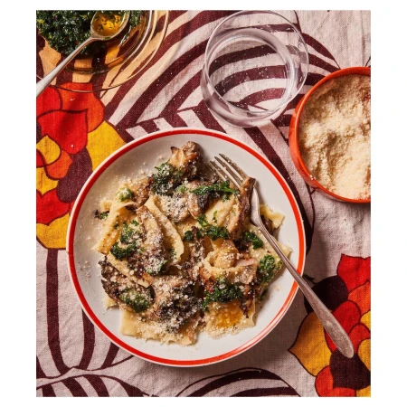 Overhead shot of a plate filled with pasta, mushrooms, and greens, alongside a glass of water and a bowl of cheese.