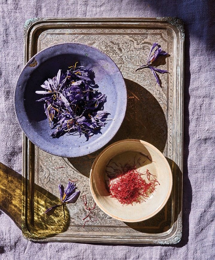 Overhead shot of saffron threads in a cream bowl and purple flower petals in a violet bowl on an ornate silver tray with a lavender linen background.