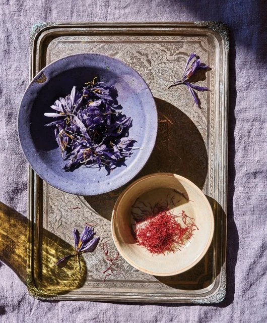 Overhead shot of saffron threads in a cream bowl and purple flower petals in a violet bowl on an ornate silver tray with a lavender linen background.