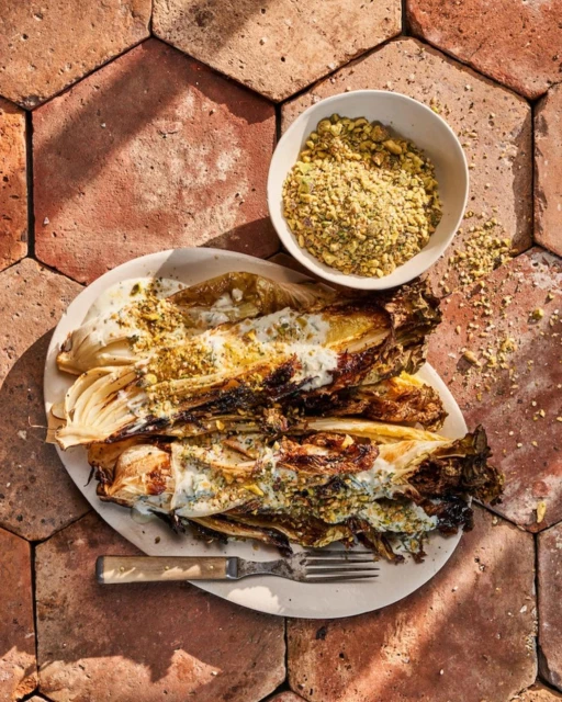 Overhead food photography of charred endives with sauce and pistachios on a plate, next to bowl of pistachios on terracotta tiles.