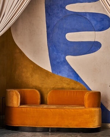 Indoor shot of a retro-style orange velvet loveseat against a white wall with blue and gold abstract designs and white curtains.