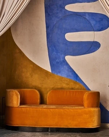 Indoor shot of a retro-style orange velvet loveseat against a white wall with blue and gold abstract designs and white curtains.