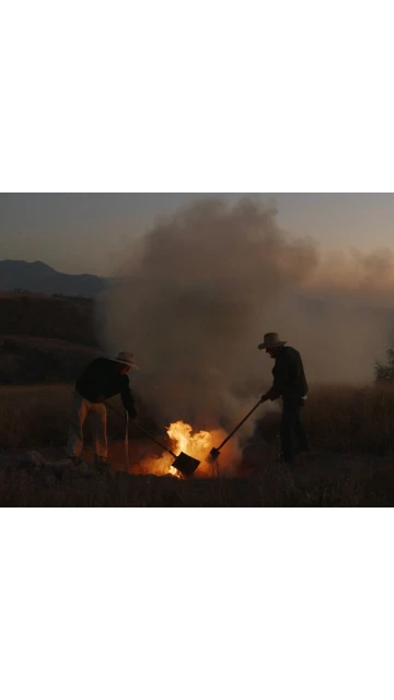 Two people wearing hats tend a fire in a rural landscape at dusk, with smoke rising against the backdrop of distant mountains.