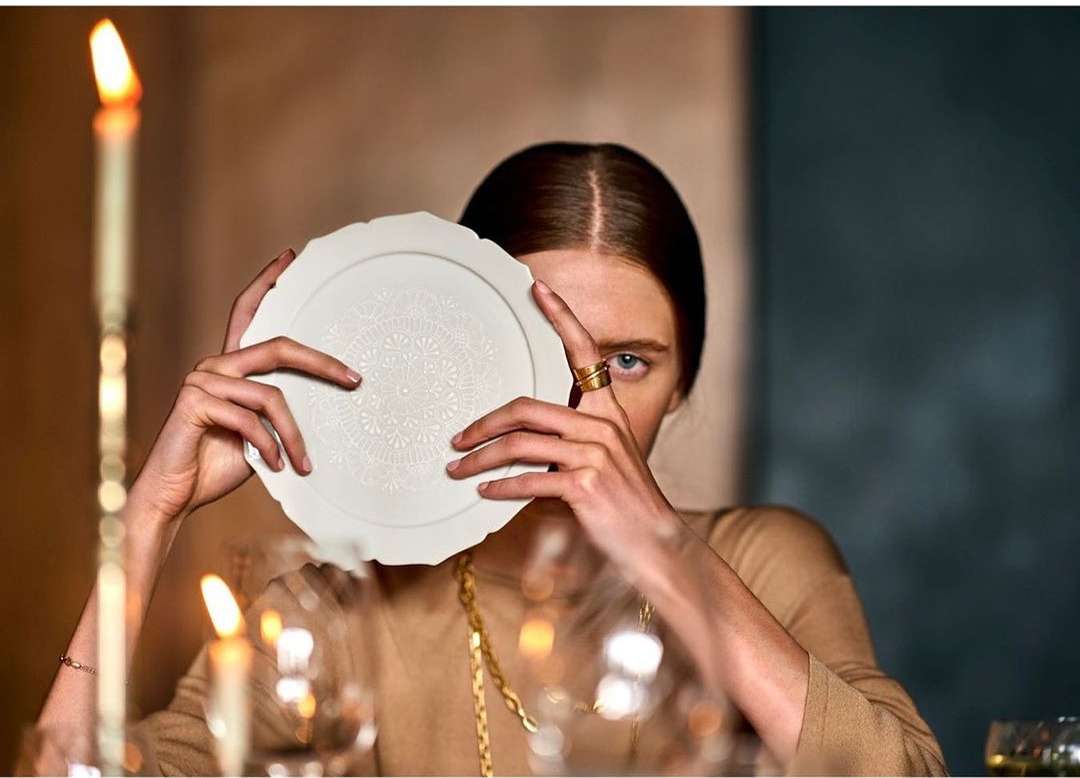 Woman holding decorative plate, partially obscuring face; lit candles and glassware on table; portrait orientation, soft focus.
