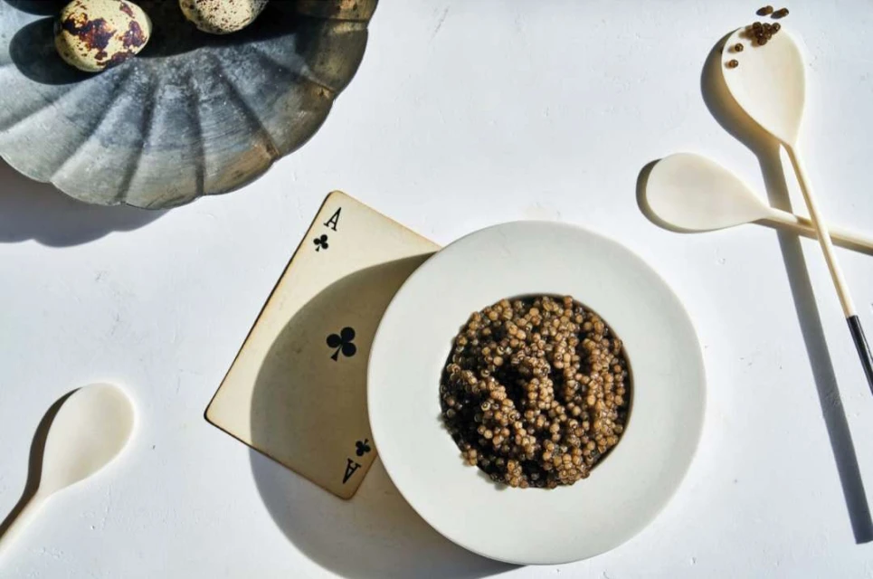 Overhead shot of caviar served in a white bowl, alongside wooden spoons, quail eggs on a metal dish, and a playing card.