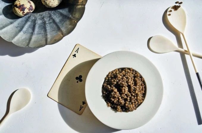 Overhead shot of caviar served in a white bowl, alongside wooden spoons, quail eggs on a metal dish, and a playing card.