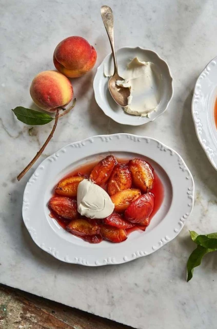 Overhead shot of a white plate with baked peaches, a dollop of cream, and fresh peaches on a marble surface.