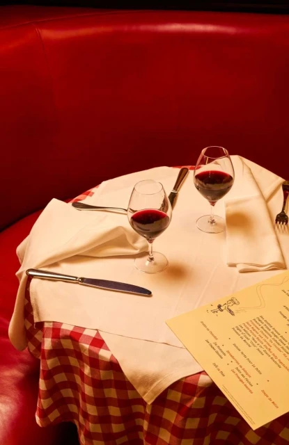 Two glasses of red wine sit on a table covered with a white and red checkered tablecloth, with a menu next to the glasses.