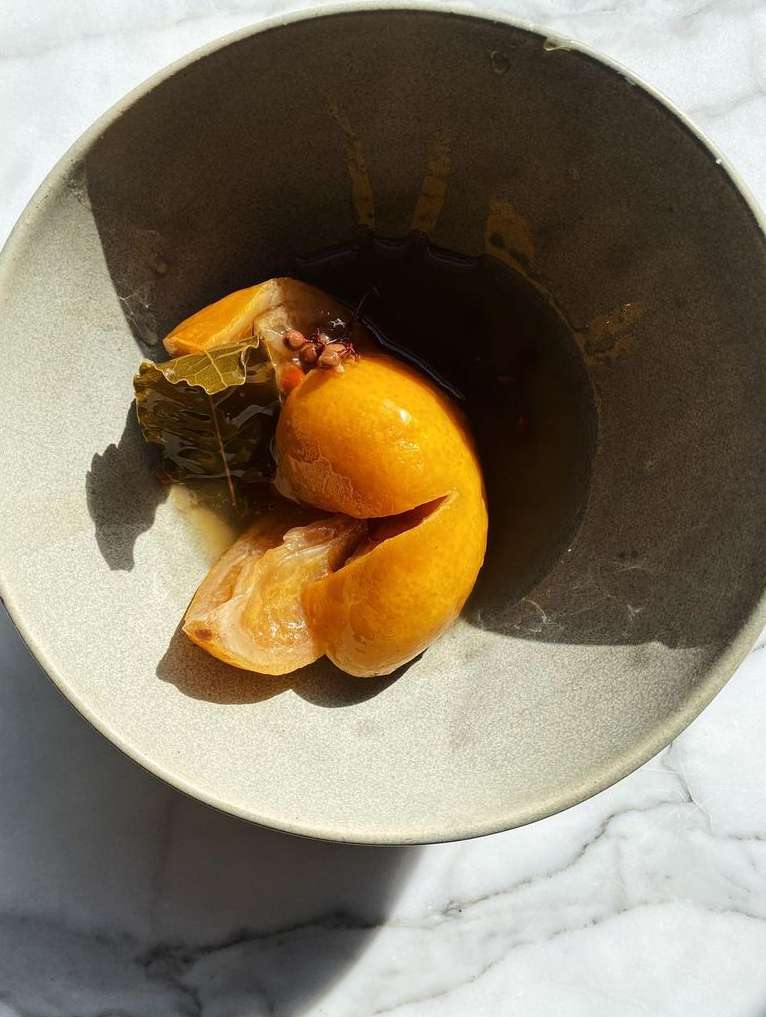 A halved preserved lemon with bay leaves and spices in a dark ceramic bowl on a marble surface.